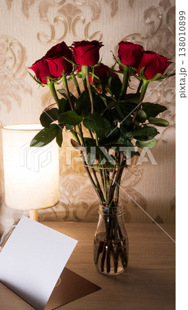 Vertical photo of a bouquet of red roses in a glass jar on a wooden table next to a glowing lamp. Blank white greeting card mockup with copy space for text. Romantic Valentine's Day gift concept Vertical photo of a bouquet of red roses in a glass jar on a wooden table next to a glowing lamp. Blank white greeting card mockup with copy space for text. Romantic Valentine's Day gift concept 138010899