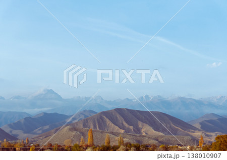 Vast mountain range landscape with rolling dry hills and autumn trees. Layered peaks fading into atmospheric haze under a clear blue sky. Copy space for text Vast mountain range landscape with rolling dry hills and autumn trees. Layered peaks fading into atmospheric haze under a clear blue sky. Copy space for text 138010907