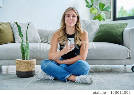 Smiling young woman with smartphone looking at camera sitting at home 138011005