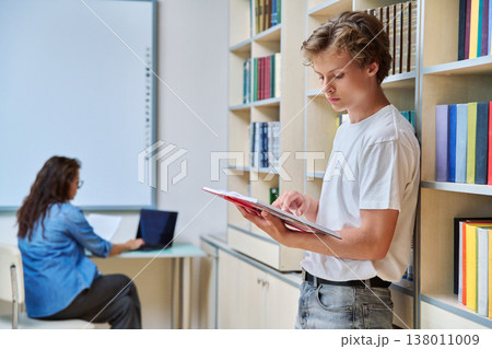 Portrait of handsome teenage boy reading book in library 138011009