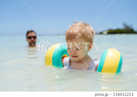 Toddler girl swimming in sea with arm floaties while father in background. 138012223
