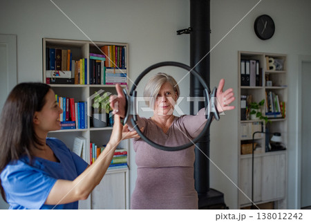 Senior woman performing physiotherapy exercise with ring under physiotherapist supervision at home. Senior woman performing physiotherapy exercise with ring under physiotherapist supervision at home. 138012242