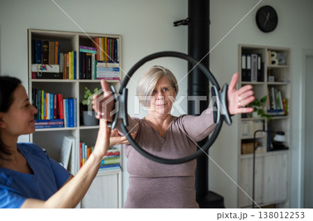 Senior woman performing physiotherapy exercise with ring under physiotherapist supervision at home. 138012253