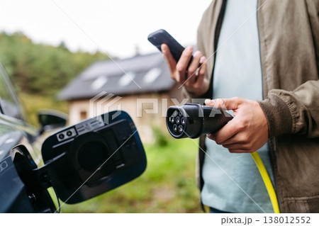 Man charging electric car in front of his house, plugging the charger into the charging port. House with solar panel system on roof behind him. 138012552
