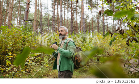 Handsome man watching wildlife with binoculars, enjoying peaceful atmosphere of forest, forestbathing Handsome man watching wildlife with binoculars, enjoying peaceful atmosphere of forest, forestbathing 138012562