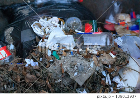Household waste, glass bottles, and cigarette butts piled on dry leaves inside a dumpster. Close-up view highlighting 138012937