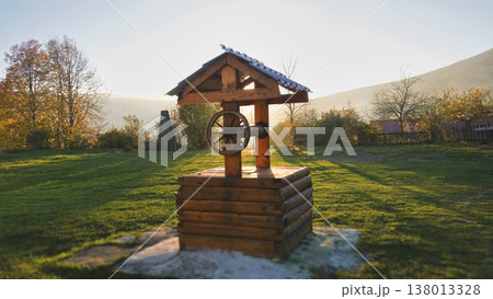 Traditional wooden water well with a small roof standing in a sunny rural garden landscape 138013328