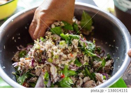 Hand mixing spicy Thai mackerel salad with herbs shallot and chili in bowl showing fresh ingredients bold flavor texture and authentic street food preparation 138014131