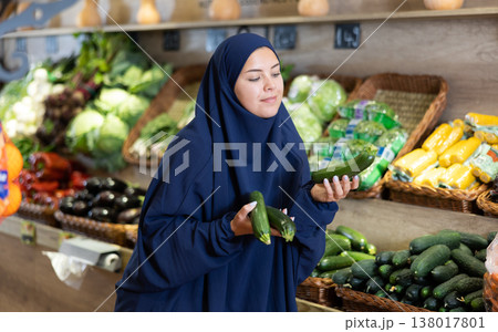 Delighted young Muslim woman purchaser choosing cucumber in grocery store 138017801