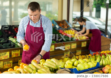 Male seller working in supermarket and lays out oranges on counter 138017886