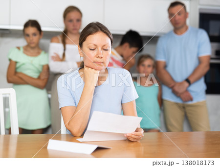 Upset mum is sitting in kitchen, reading letter. Blurred worried family in background Upset mum is sitting in kitchen, reading letter. Blurred worried family in background 138018173