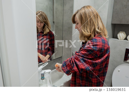 Woman cleaning bathroom sink and mirror with cloth, focused on wiping dust from faucet and vanity, plaid shirt, reflective mirror showing profile, tidy contemporary bathroom, soft natural light, calm 138019631
