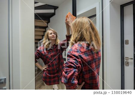 caucasian woman dusting mirror in hallway with plaid shirt and soft smile, wiping reflection and wood trim, bright modern staircase visible behind, casual morning routine showing tidy domestic upkeep 138019640
