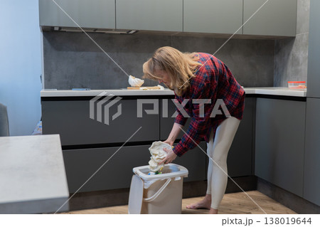 Blonde white woman emptying bowl into trash bin in tidy modern kitchen, performing cleanup after baking. Casual stance with one hand on counter and other tipping bowl shows everyday household chore. 138019644