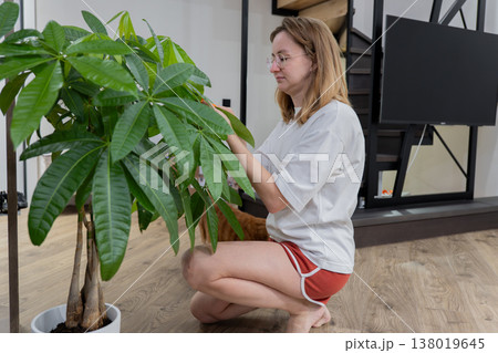 woman wiping dusty plant leaves gently kneeled on wood floor in modern living area, casual shorts and white blouse using soft cloth to remove dust from large potted foliage, warm natural light, 138019645