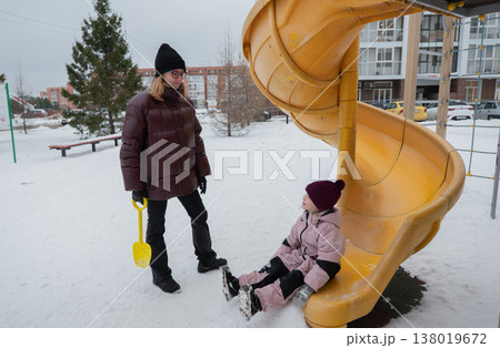 caucasian mother supervising daughter on snowy playground slide, holding yellow shovel, bundled puffer coats, knit hats, boots gentle smile, watching play, visible breath, apartment buildings caucasian mother supervising daughter on snowy playground slide, holding yellow shovel, bundled puffer coats, knit hats, boots gentle smile, watching play, visible breath, apartment buildings 138019672