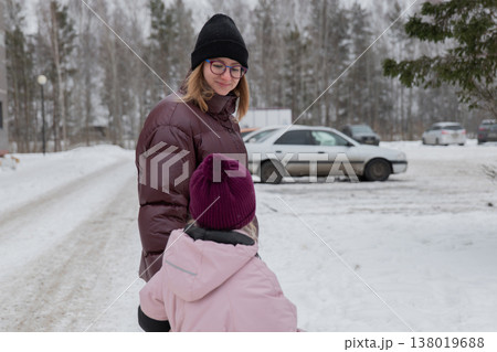 Volunteer guiding child in snowy parking, parked cars and forest backdrop, adult wearing glasses and puffer coat, child in pink jacket and purple beanie, careful steps on salted asphalt, casual 138019688