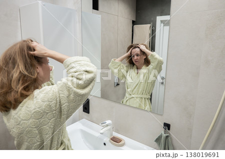Caucasian woman at mirror checking expression while running hands through hair in bathrobe, serious concentrated look, modern bathroom sink and shower visible, preparing for audition with tense 138019691