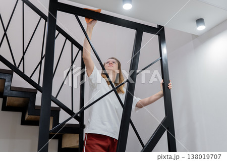 Young woman dusting staircase railing, reaching upward, wearing white shirt and red skirt, modern interior, focused expression, soft ceiling lights, tidy minimalist hallway, domestic chore, homemaker 138019707