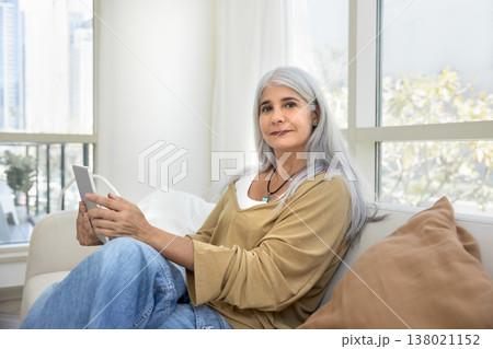 Mature female sits on couch with tablet, looking at camera 138021152