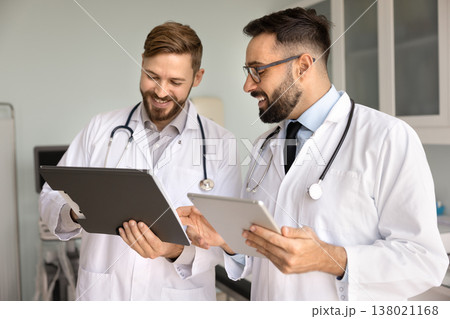 Friendly smiling male doctors discuss diagnosis using paper electronic documents Friendly smiling male doctors discuss diagnosis using paper electronic documents 138021168