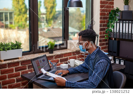 Male analyst with clipboard, researching financial indicators on laptop and planning business presentation. Black man seated in brick wall office is comparing company strategy ideas with documents. Male analyst with clipboard, researching financial indicators on laptop and planning business presentation. Black man seated in brick wall office is comparing company strategy ideas with documents. 138021252