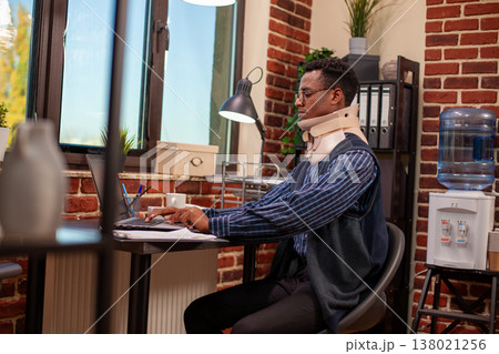 Entrepreneur wearing neck brace works on his laptop in startup office, recovering from injury and focusing on his business project. Black businessman concentrating on company research after accident. Entrepreneur wearing neck brace works on his laptop in startup office, recovering from injury and focusing on his business project. Black businessman concentrating on company research after accident. 138021256