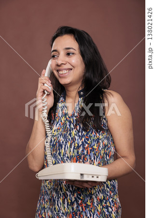 Smiling indian woman answering landline phone call, customer support service, cheerful consultant. Lady chatting, person with positive facial expression holding retro telephone Smiling indian woman answering landline phone call, customer support service, cheerful consultant. Lady chatting, person with positive facial expression holding retro telephone 138021426