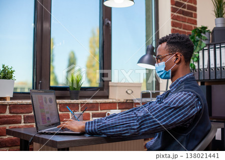 African american manager wearing earbuds, typing on digital device and researching marketing strategy at desk. Office worker with face mask, using laptop to review projects as he listens to podcasts. 138021441