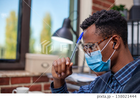 Closeup of male analyst wearing face mask and holding pen near his forehead in brick wall office. Black manager with earbuds, leaning on table and tapping his head, focused on company research project 138021444