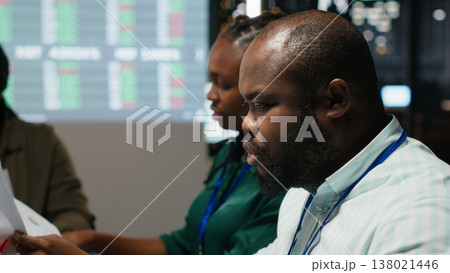 Black manager and his team studying annual data reporting during meeting late at night, looking into market supply and demands to facilitate global business development in boardroom. Camera B. 138021446