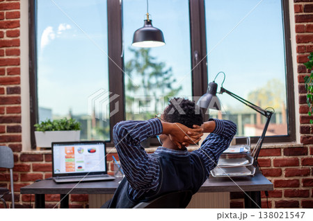 Businessman leans in office chair, hands resting behind his head, stretching and taking a break after busy schedule. African american male manager relaxing at his desk, feeling satisfied after work. Businessman leans in office chair, hands resting behind his head, stretching and taking a break after busy schedule. African american male manager relaxing at his desk, feeling satisfied after work. 138021547
