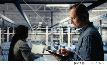 Data center admin talking in audio headset mic, helping clients resolve website hosting problems. Server farm worker on industrial platform helping users keep online services running, camera A 138021607