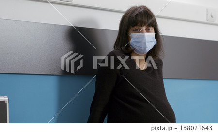 Portrait of female patient expecting baby in medical office, attending checkup appointment with health specialist. Pregnant woman sitting on bed and wearing face mask at consultation. Portrait of female patient expecting baby in medical office, attending checkup appointment with health specialist. Pregnant woman sitting on bed and wearing face mask at consultation. 138021643