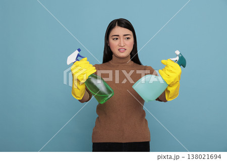 Asian young inactive dull housekeeper from chores holding two detergent spray in yellow gloves against blue background, Cleaning home concept, Unhappy amazed with a negative state of mind, Asian young inactive dull housekeeper from chores holding two detergent spray in yellow gloves against blue background, Cleaning home concept, Unhappy amazed with a negative state of mind, 138021694