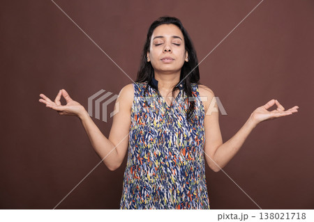 Serene indian woman meditating, showing om sign with fingers, front view studio mid shot on brown background. Relaxed person with closed eyes, calm lady with yoga symbol, meditation gesture Serene indian woman meditating, showing om sign with fingers, front view studio mid shot on brown background. Relaxed person with closed eyes, calm lady with yoga symbol, meditation gesture 138021718