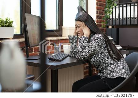 Frustrated female manager leaning on table, overwhelmed by marketing strategy preparation on computer. Asian businesswoman holds forehead in hands at desk, experiencing a headache in modern workspace. Frustrated female manager leaning on table, overwhelmed by marketing strategy preparation on computer. Asian businesswoman holds forehead in hands at desk, experiencing a headache in modern workspace. 138021787