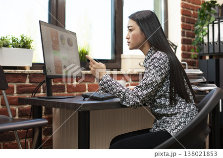 Female analyst pointing at computer screen, closely reviewing financial graphs in startup office. Young businesswoman concentrating on her desktop monitor, looking at company research information. 138021853