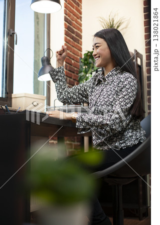 Asian woman celebrates at desk with her fist raised, after reading positive business outcomes on her computer. Excited female manager smiles, reflecting on success of company project. Asian woman celebrates at desk with her fist raised, after reading positive business outcomes on her computer. Excited female manager smiles, reflecting on success of company project. 138021864