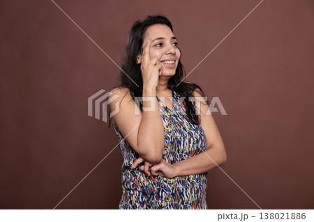 Smiling woman laughing, looking upwards portrait, model holding finger on face. Pensive cheerful person standing, happy lady thinking, studio medium shot on brown background 138021886