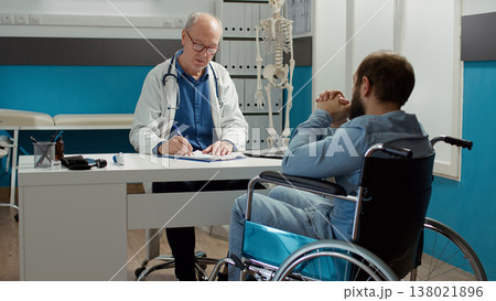 Health specialist doing consultation with wheelchair user in disability friendly cabinet. Medic examining patient with health condition and impairment, using expertise to talk to paralyzed man. Health specialist doing consultation with wheelchair user in disability friendly cabinet. Medic examining patient with health condition and impairment, using expertise to talk to paralyzed man. 138021896