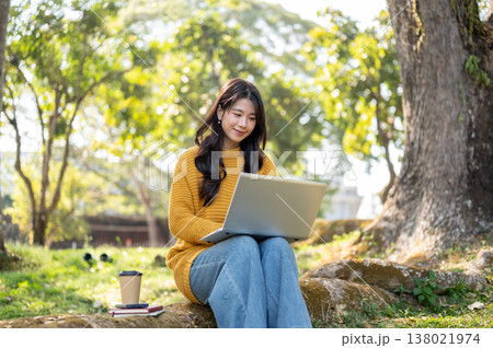 Pretty asian woman in yellow sweater looking at laptop while sits on tree root with book and coffee. 138021974