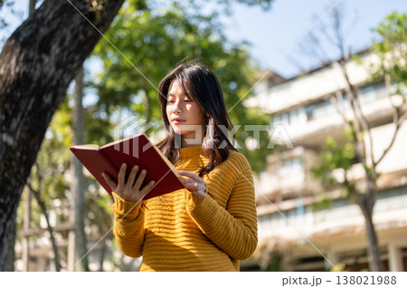 Pretty asian woman in yellow sweater holding and reading a book while standing under tree in park. 138021988