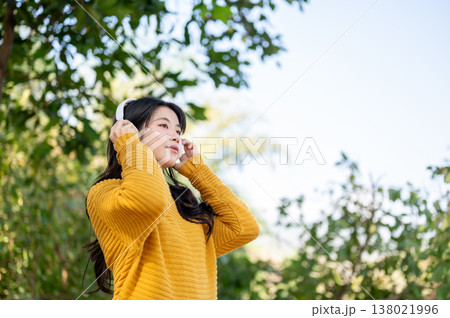 Pretty asian woman in yellow sweater wearing headphones listening to music while standing in park. Pretty asian woman in yellow sweater wearing headphones listening to music while standing in park. 138021996
