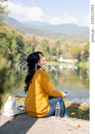 Happy asian woman holding phone and sitting with bag and water bottle on sidewalk aside river lake. Happy asian woman holding phone and sitting with bag and water bottle on sidewalk aside river lake. 138022003