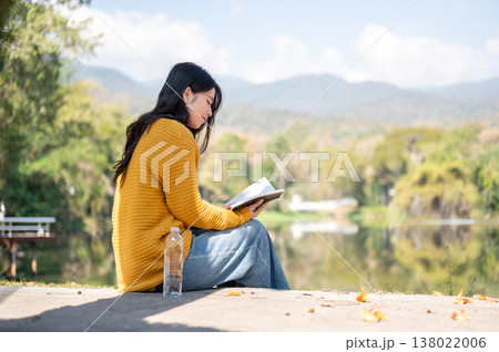Asian woman reading a book while sits with water bottle on sidewalk aside river lake under sunlight. 138022006