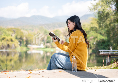 Asian woman reading book while sitting with water bottle on sidewalk aside river lake under sunlight 138022007