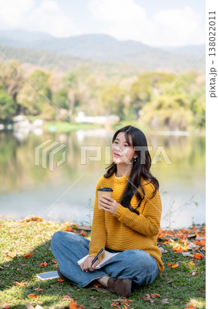 Pretty asian woman holding drinking coffee while sitting crossed legs on grass lawn aside river lake Pretty asian woman holding drinking coffee while sitting crossed legs on grass lawn aside river lake 138022011