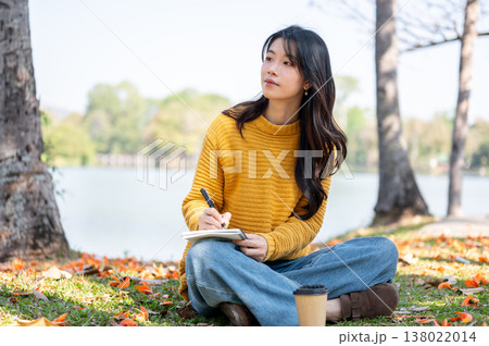 Pretty asian woman holding pen writing diary over crossed legs as sit on grass lawn aside river lake Pretty asian woman holding pen writing diary over crossed legs as sit on grass lawn aside river lake 138022014