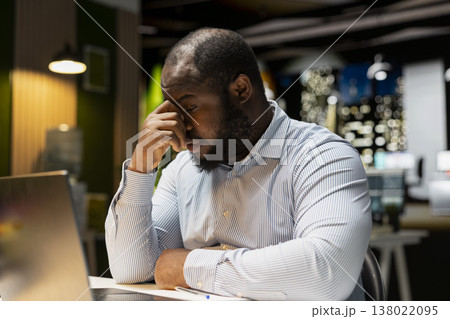 African american weary man struggling to stay awake after hours in the office, trying hard to work and finish task. Person feeling overworked and overwhelmed after hours, sleep deprivation. African american weary man struggling to stay awake after hours in the office, trying hard to work and finish task. Person feeling overworked and overwhelmed after hours, sleep deprivation. 138022095